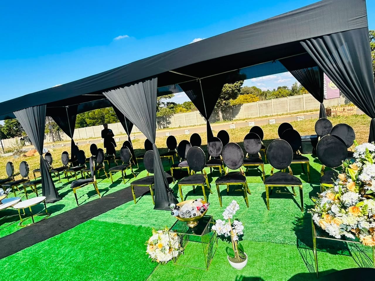 Black-draped funeral tent with black-and-gold chairs and floral tributes